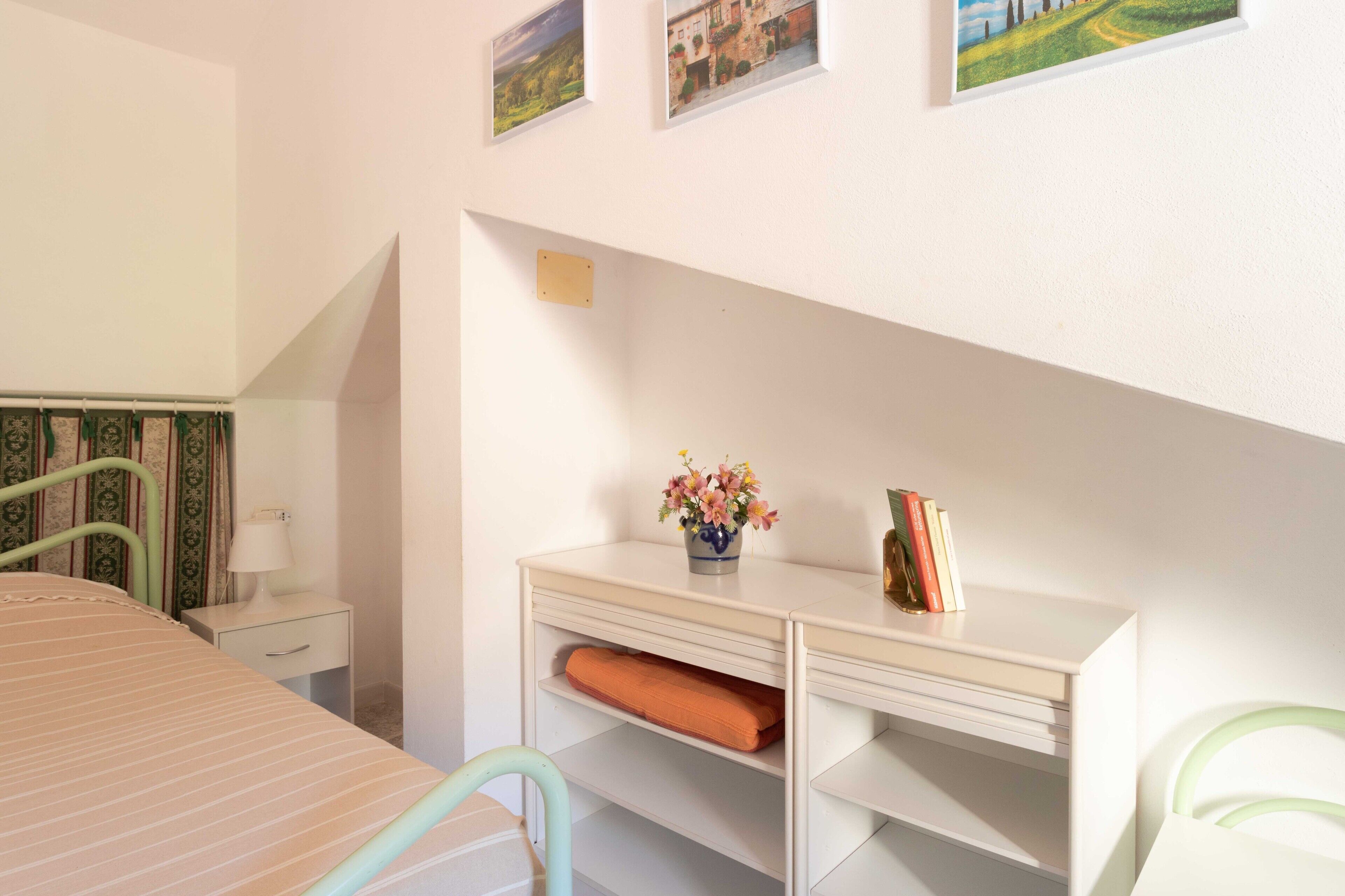 A bedroom with a bed, a desk with a lamp, a chair, and a shelf with books and a vase of flowers.