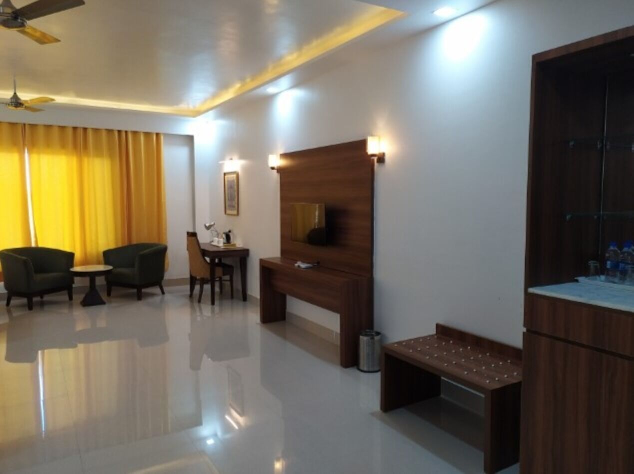 A modern hotel room with a flat-screen TV, a wooden headboard, a small dining table, two chairs, a bench, and a minibar.