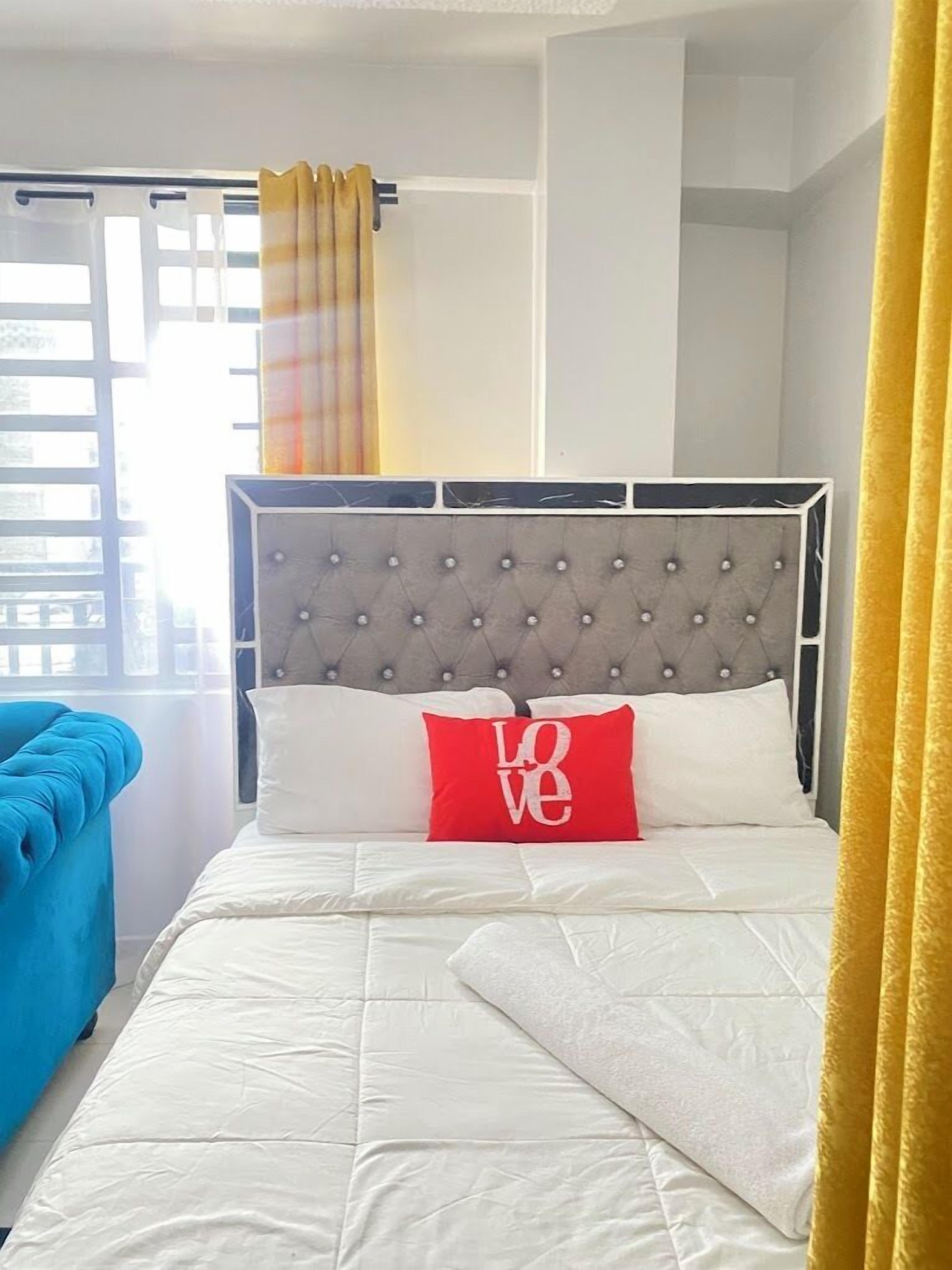 A neatly made bed with a tufted headboard, a red pillow with the word "LOVE," and a blue chair to the left.