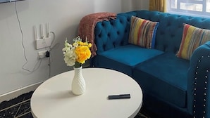 En moderne stue med en blå sofa, et rundt hvidt sofabord med en vase blomster, et fladskærms-tv monteret på væggen og et vindue med gule gardiner.