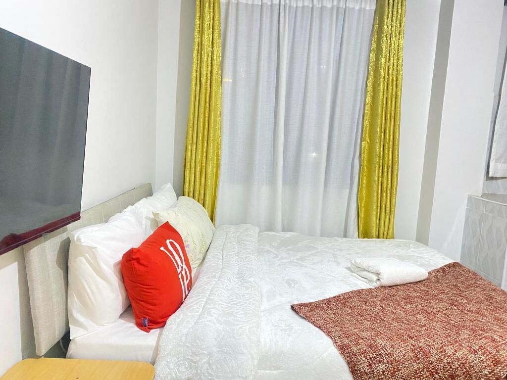 Una habitación de hotel con cama, ropa de cama blanca, almohada roja y cortinas amarillas.