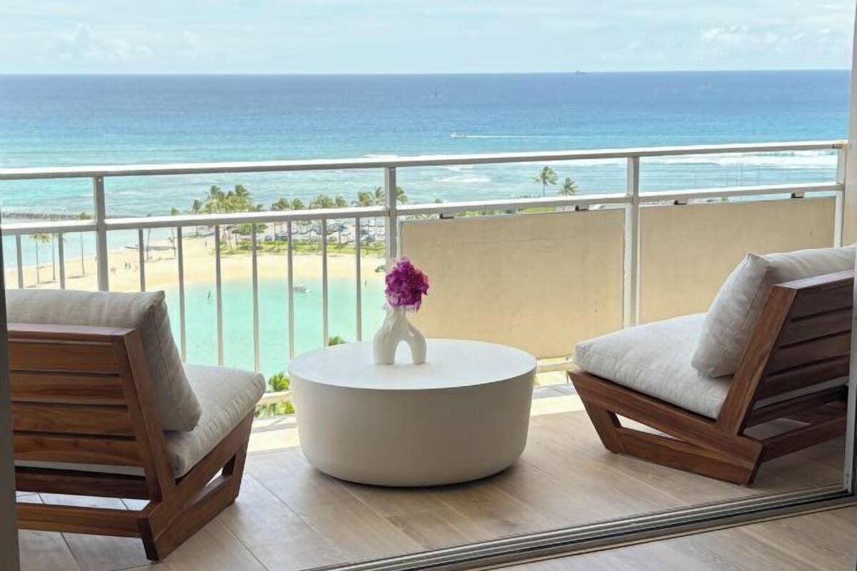 A balcony with a view of the ocean, two wooden chairs, a white round table, and a vase with purple flowers.
