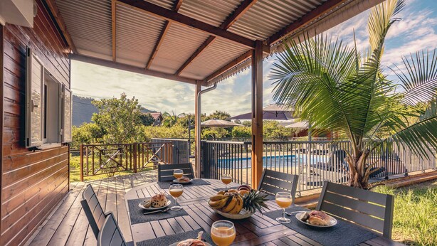 Un área de comedor al aire libre con mesas y sillas de madera, y una piscina al fondo.
