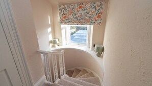 Un escalier avec une plateforme intermédiaire incurvée, une fenêtre agrémentée d’une lambrequin à motifs floraux et deux lampes fixées au mur.