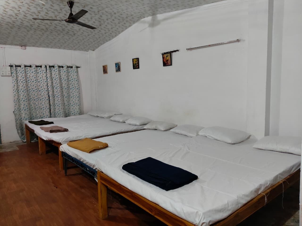 A dormitory room with two single beds, wooden frames, white bedding, a ceiling fan, and a curtain with a floral pattern.