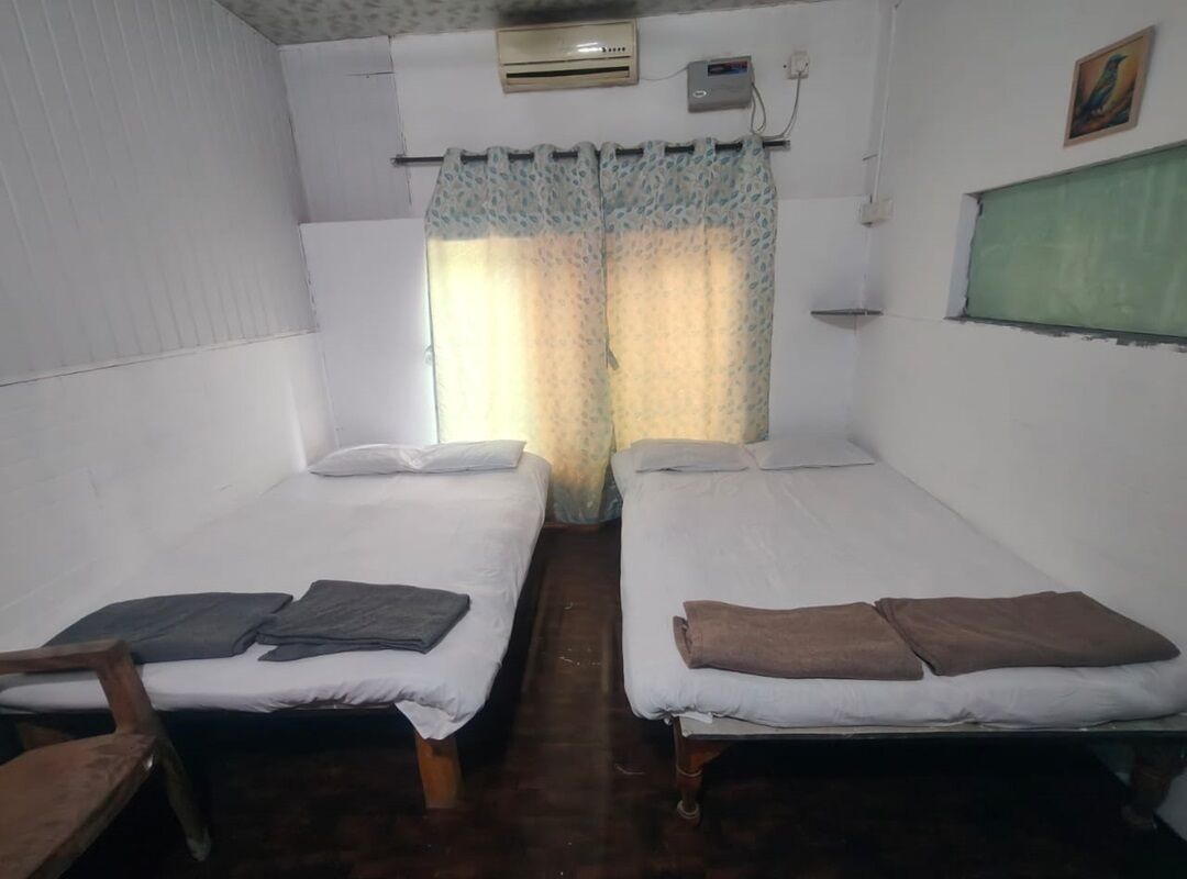 A room with two single beds, a wooden chair, a small table, a window with a green blind, and an air conditioning unit.
