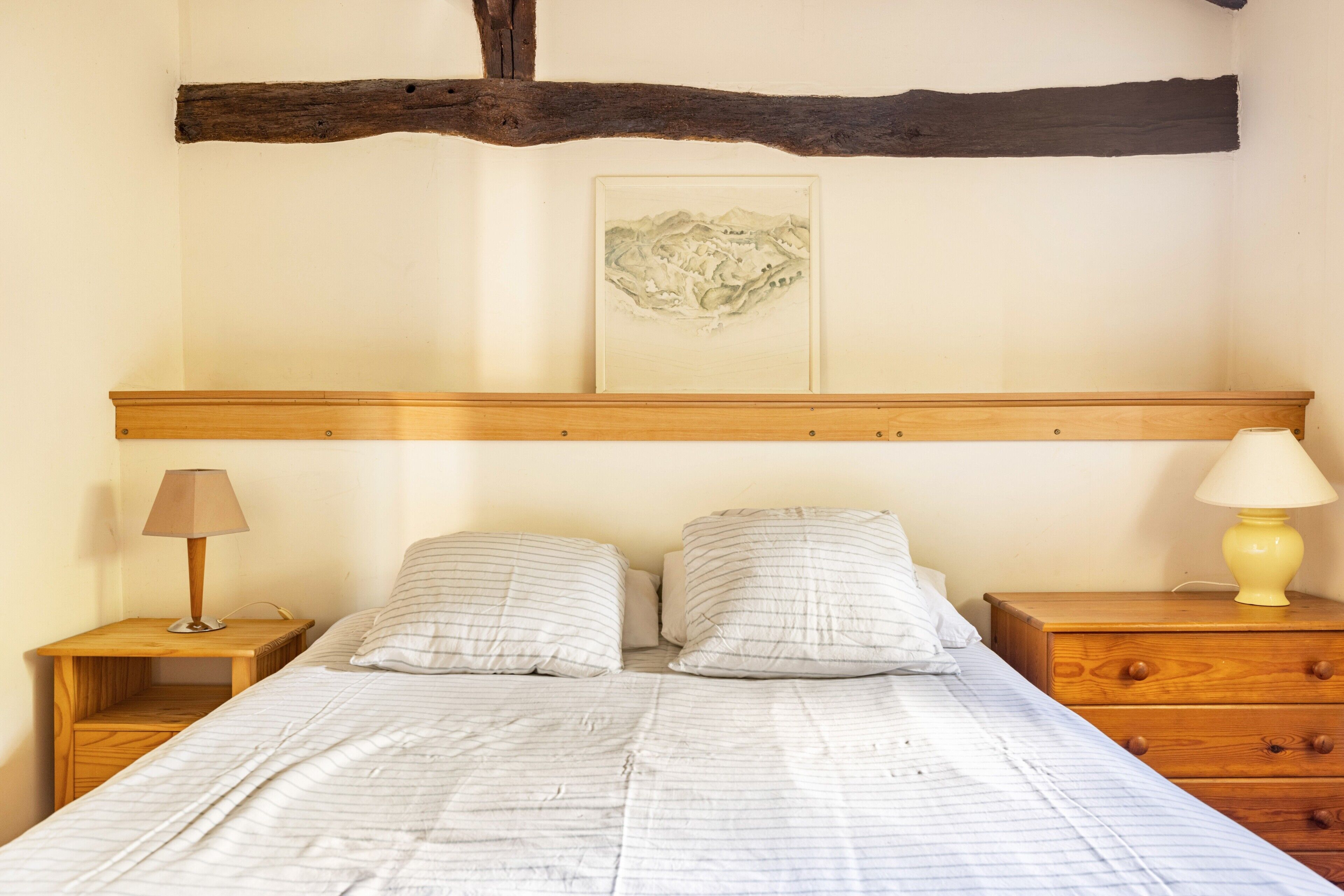 Een slaapkamer met een houten hoofdbord, een bed met gestreept beddengoed, twee nachtkastjes met lampen, en een ingelijst schilderij aan de muur.