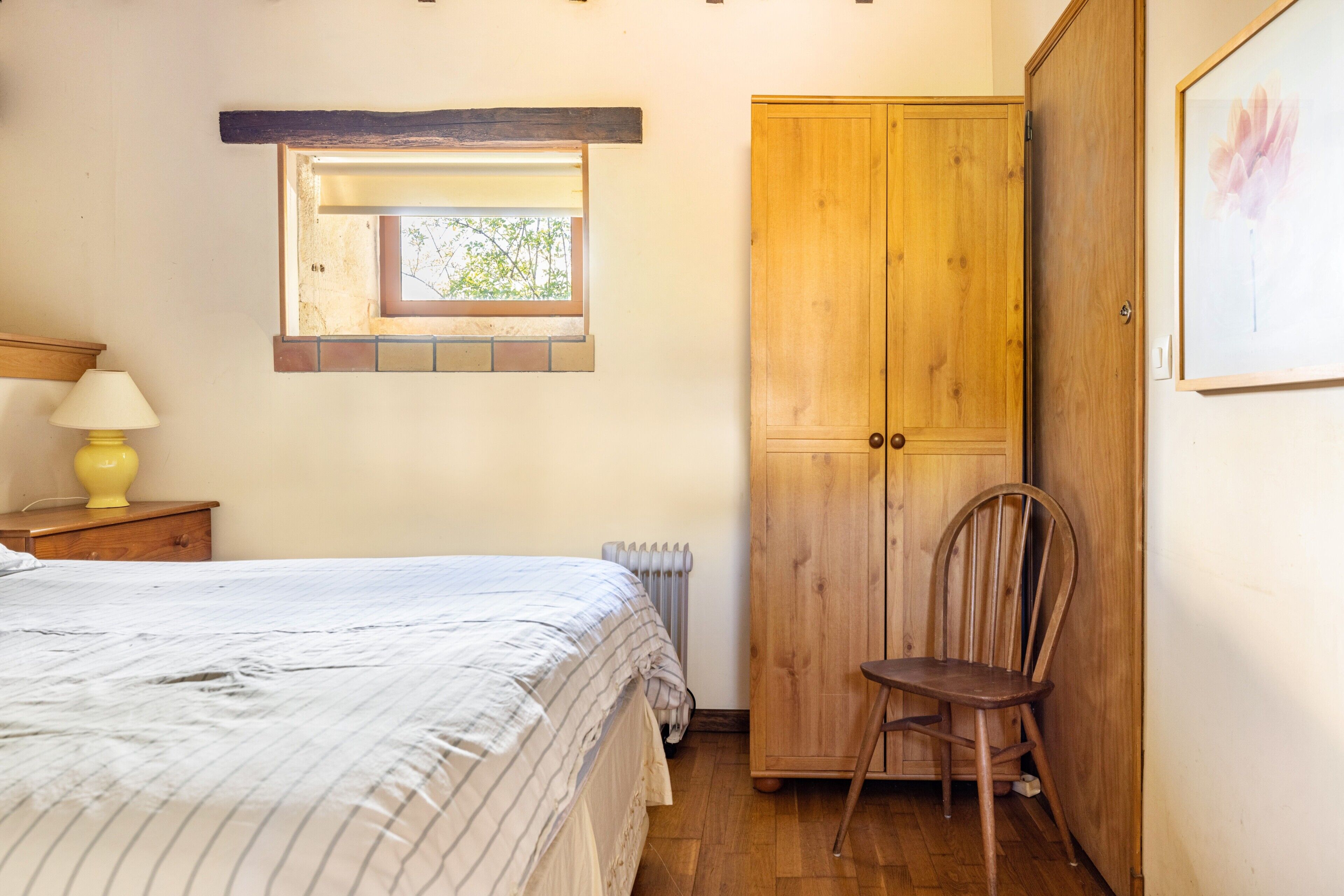 Un dormitorio con cama, un armario de madera, una silla, una mesita de noche con lámpara y una ventana con vista a árboles.