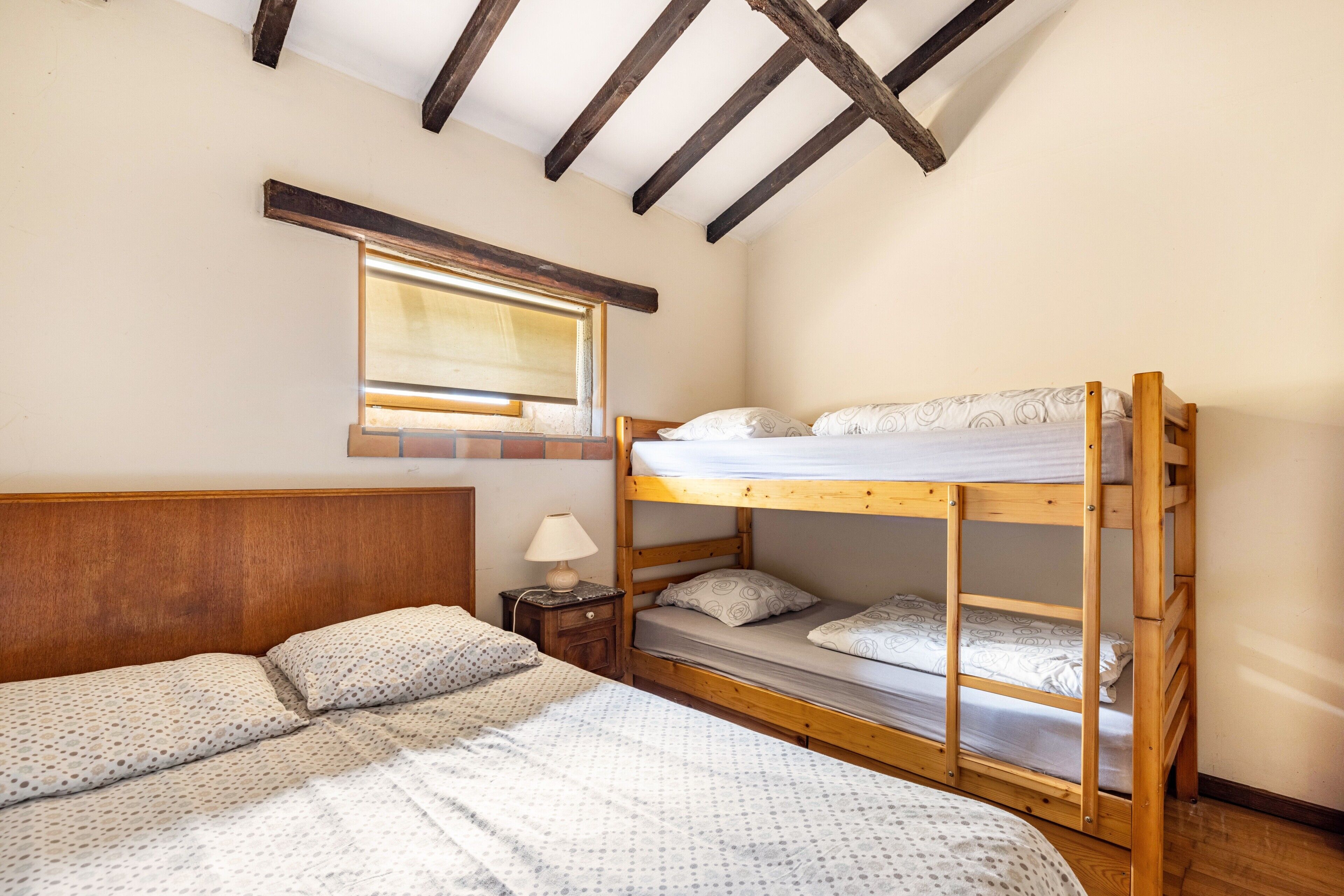A room with a bunk bed, a single bed, a nightstand with a lamp, a window with blinds, and wooden beams on the ceiling.