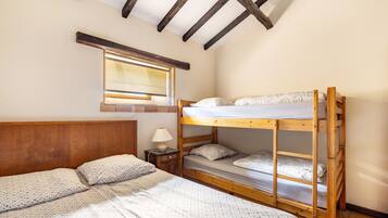 A room with a bunk bed, a single bed, a nightstand with a lamp, a window with blinds, and wooden beams on the ceiling.