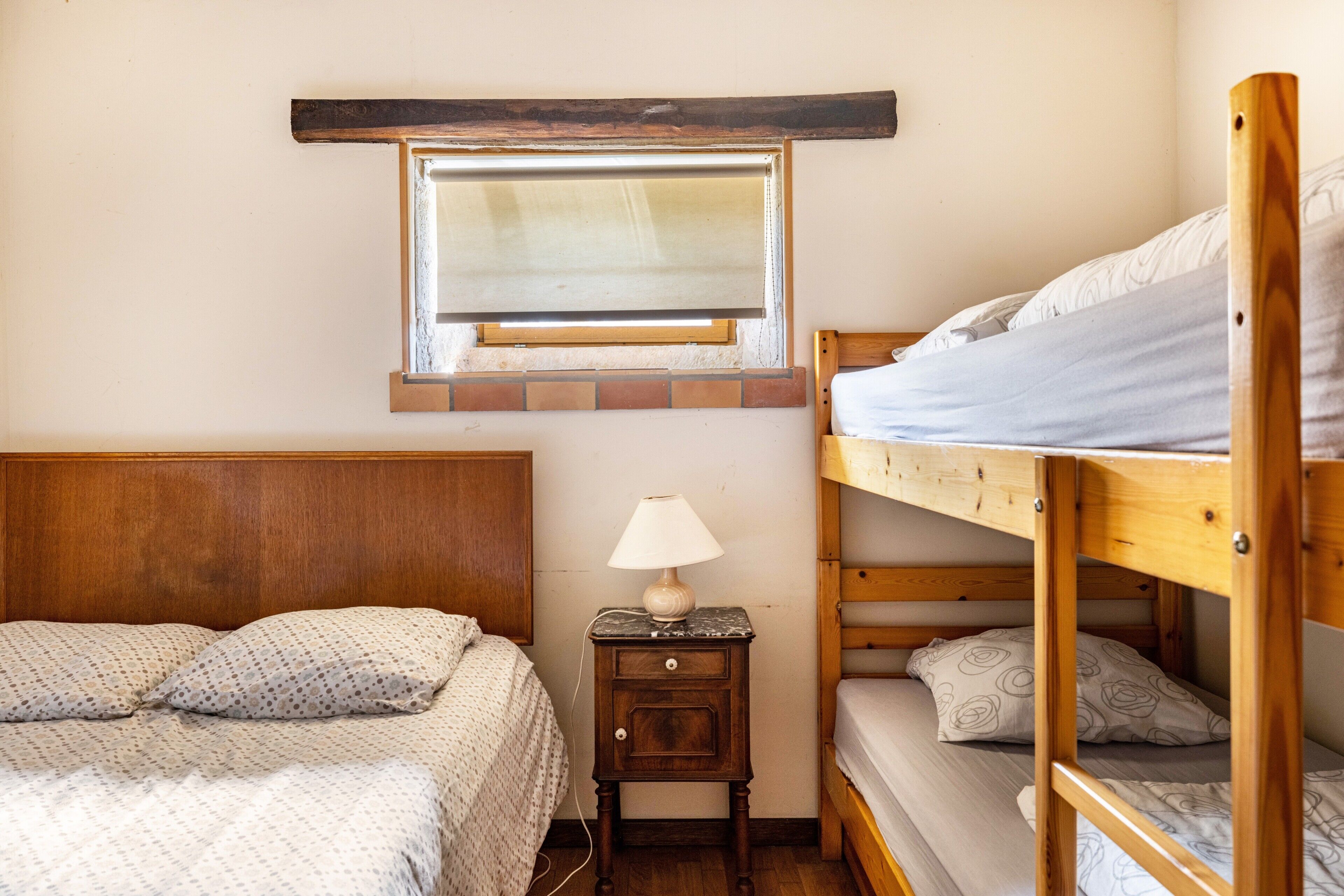 Un lit superposé avec une tête de lit en bois, un lit simple avec une couvre-lit à motifs, une table de chevet avec une lampe et une fenêtre avec un cadre en bois.