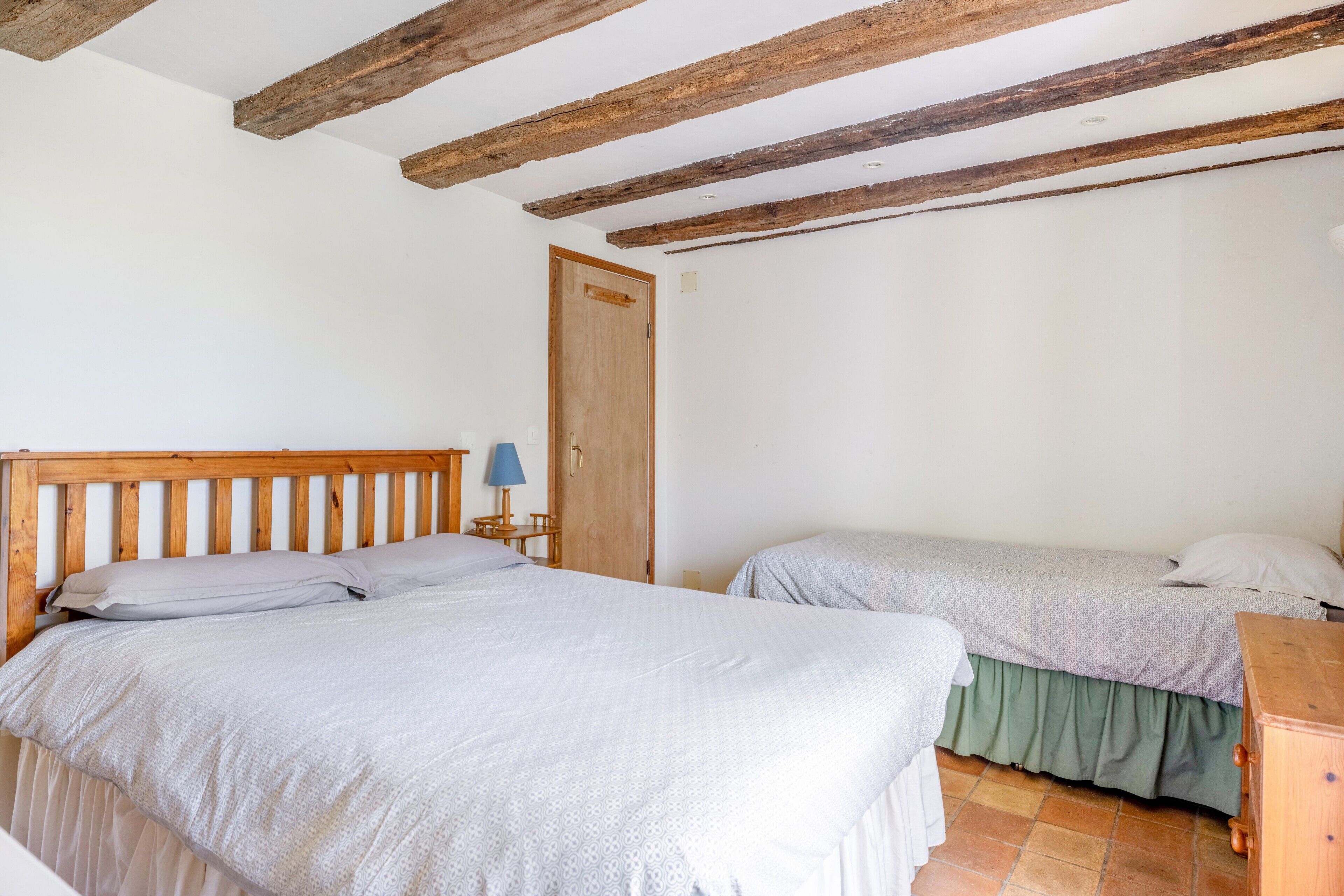 A bedroom with two beds, wooden beams on the ceiling, a wooden headboard, a wooden nightstand with a lamp, and a wooden dresser.