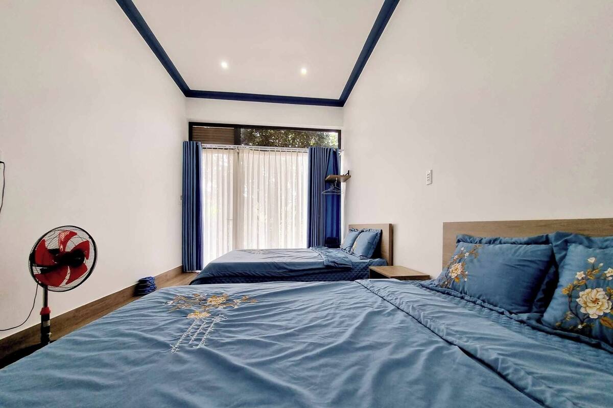 Une chambre avec un grand lit, une table de chevet, un ventilateur et une fenêtre avec des rideaux.