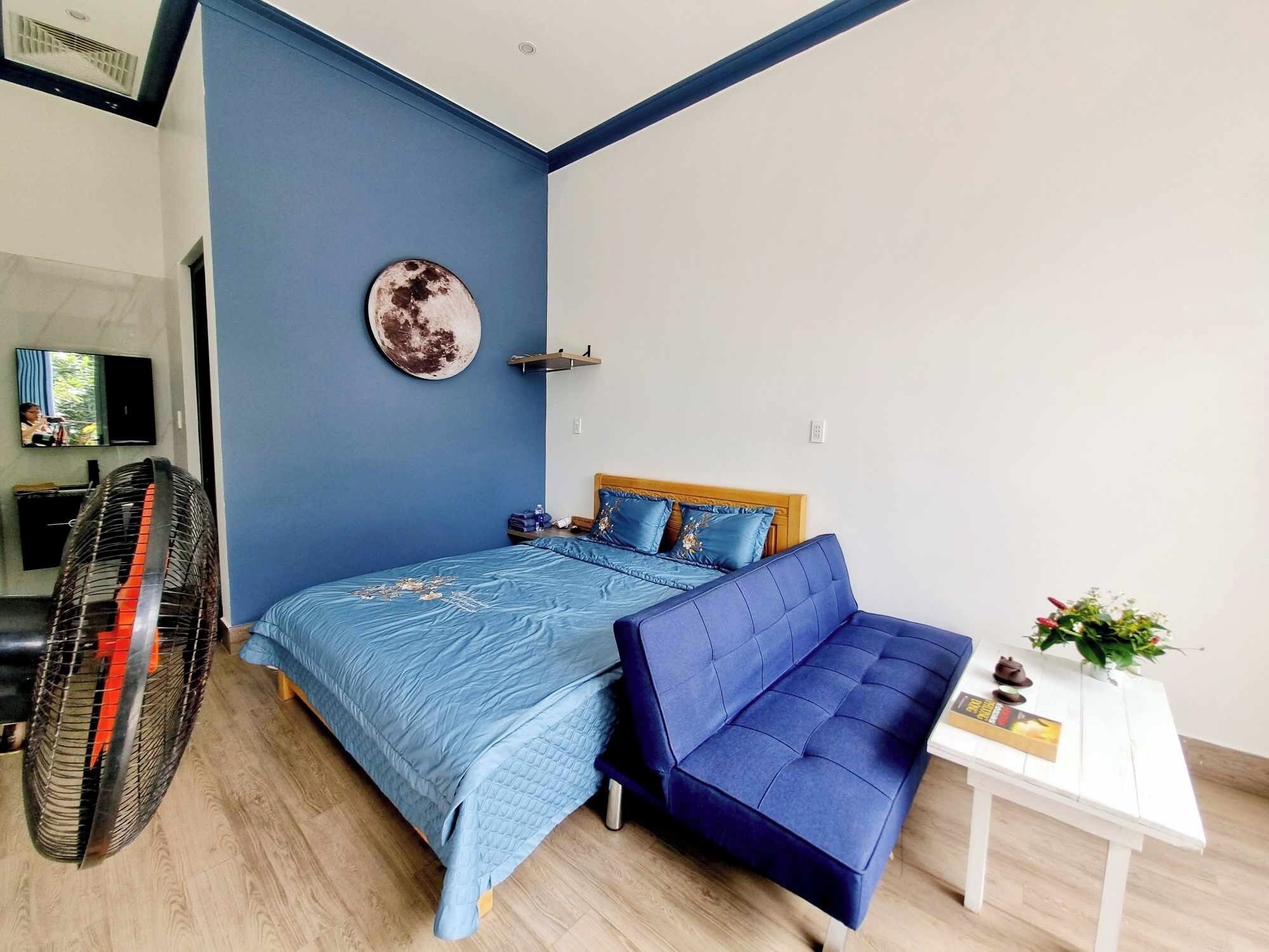 A bedroom with a bed, a blue sofa, a small table with a plant, and a wall-mounted fan.