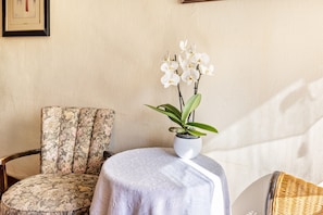 Ett rum med en stol med blommönster, ett litet runt bord med ett vitt tyg över och en orkidé i en kruka.