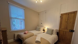 A room with two beds, a window with blinds, a radiator, a wooden door, and a small table with a lamp. - City Centre, River views, Sleeps 5, Free Parking (Canterbury)