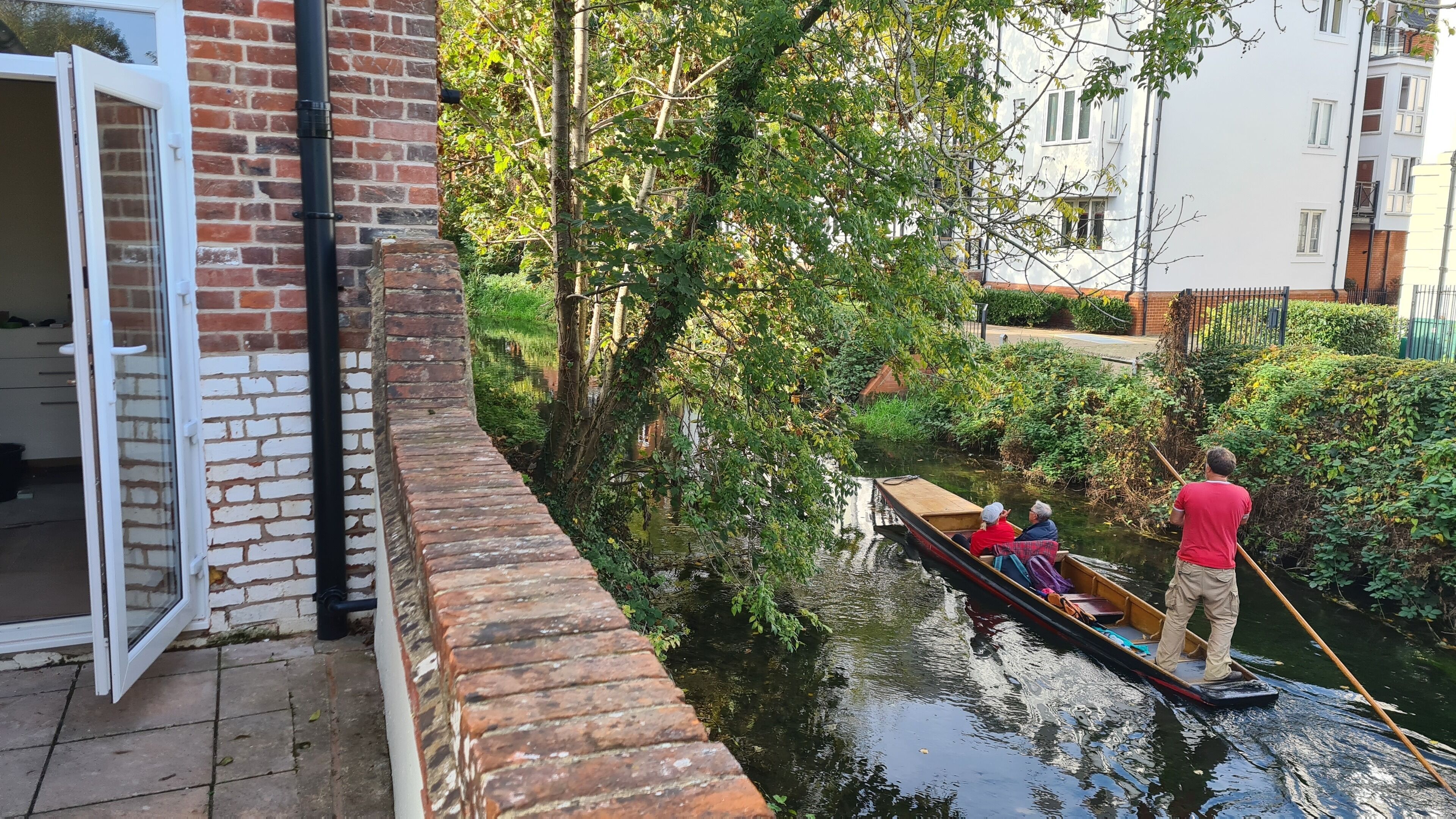 A narrow canal with a rowboat carrying two people and a man standing with a pole.