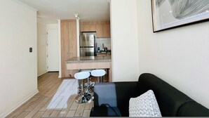 Une cuisine moderne avec des meubles en bois et des appareils en acier inoxydable, un coin repas avec un canapé noir et des tabourets de bar blancs, et une œuvre d’art encadrée au mur.