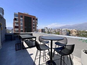 Una terraza en la azotea con una mesa, sillas, un banco y vistas a un paisaje urbano con montañas al fondo.