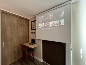 A small room with a wooden desk, a chair, and a projector screen displaying a Netflix interface. - Urbano Art Stico Irarr Zaval A C Laundry Parking (Santiago)