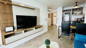 Una sala de estar moderna con televisor de pantalla plana, una mesa de centro con un objeto decorativo, un sofá azul y un comedor con sillas y mesa.