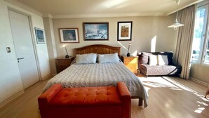 Un dormitorio con cama, una banca, mesita de noche, lámpara y cuadros en la pared.