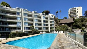 Una piscina con sillas de descanso, un edificio de varios pisos con balcones y un cielo azul despejado.