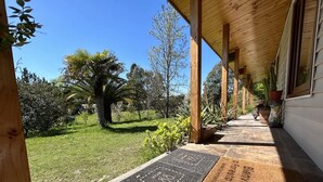 Eine überdachte Veranda mit Steinboden, Holzsäulen und Blick auf einen Garten mit Rasen und Palmen.