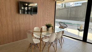 A modern dining area with a glass table and white chairs, a television mounted on a wooden wall, and a view of a balcony with outdoor seating. - Estudio Dubl Almeyda Crib Laundry Gym Pool (Santiago)