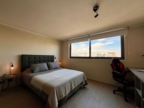 A bedroom with a bed, a desk, a chair, a window with a view of buildings, and a ceiling light. - Estudio Dubl Almeyda Crib Laundry Gym Pool (Santiago)