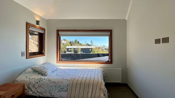 A bedroom with a bed, a window with a view of buildings, a radiator, and a small wooden nightstand. - Casa Playa Hermosa BBQ Jacuzzi Parking Wifi (Puerto Varas)