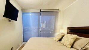 A hotel room with a bed, a TV mounted on the wall, and a sliding door leading to a balcony with a city view. - Urbano Familiar Bustamante Laundry Parking Pool (Santiago)