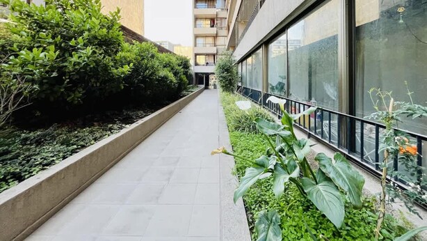 A corridor with a tiled floor, bordered by a low wall with dense green foliage and a glass building with balconies on one side. - Urbano Familiar Bustamante Laundry Parking Pool (Santiago)