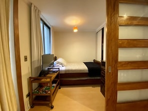 Un dormitorio compacto con una cama individual, una mesita de noche pequeña con un televisor y una ventana con cortinas.