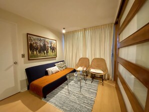 A room with a sofa, two chairs, a coffee table, a painting, and a window with curtains. - Urbano Viana Terminal Home Office Pool Wifi (Vina del Mar)