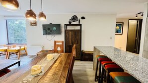 A modern kitchen with a wooden dining table, bar stools, pendant lights, and a television on the wall. - Buho Modern Nature Retreat - Puc n (Pucón)