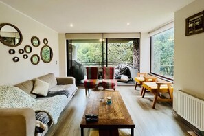 A living room with a sofa, a coffee table, and a set of chairs. There are large windows with a view of trees. - Buho Modern Nature Retreat - Puc n (Pucón)
