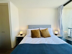 Habitación de hotel con cama, dos lámparas de noche, ventana con cortinas y una silla de balcón.