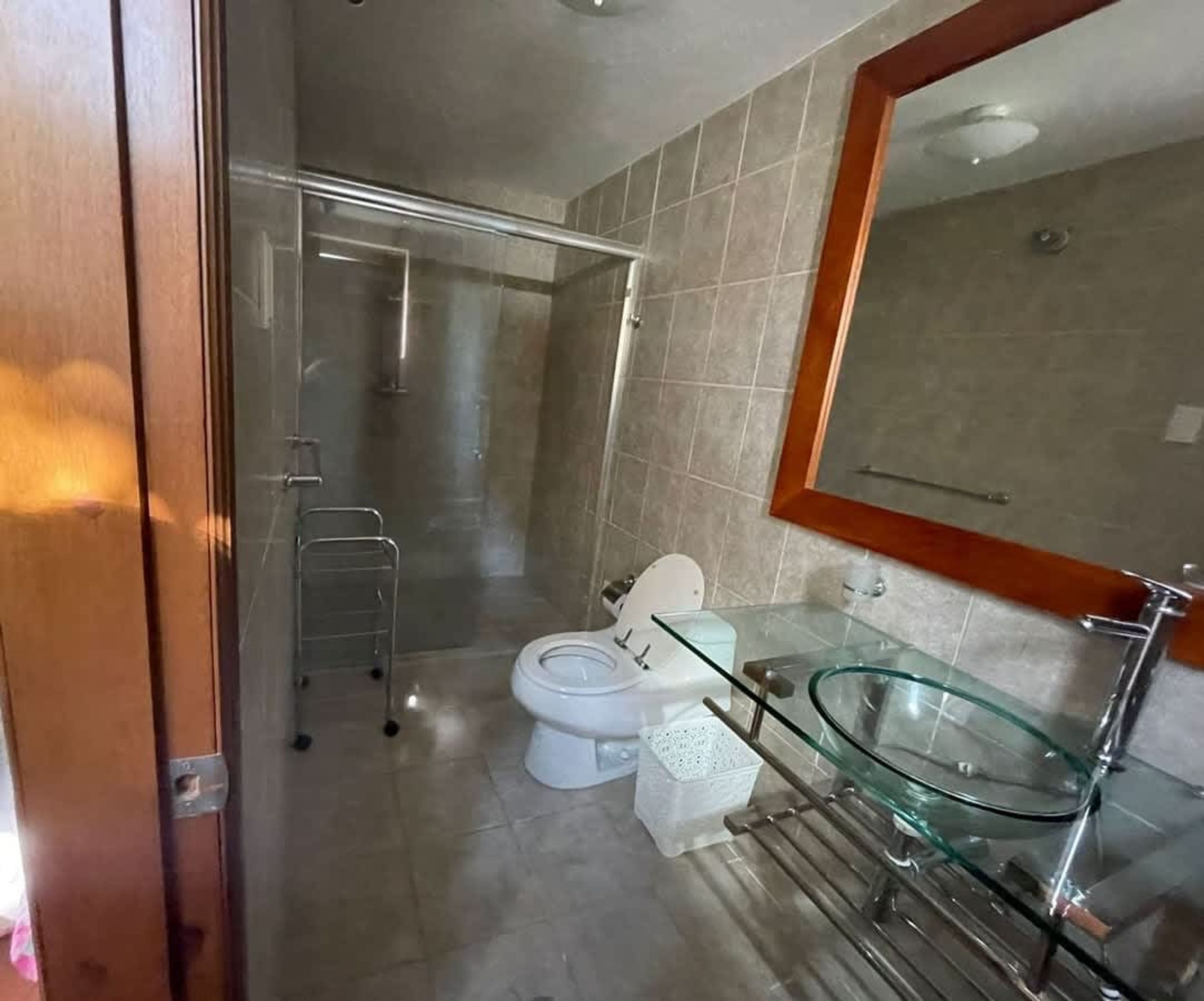 Un baño con cabina de ducha de vidrio, inodoro blanco y lavamanos de vidrio con espejo de marco de madera.