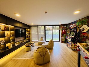 A modern living room with a flat-screen TV, comfortable seating, and a gaming area with arcade machines. - Urbano Victoria Cowork Gym Washer Wifi (Santiago)