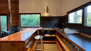 A modern kitchen with a wooden island, bar stools, a large window, and a view of the forest. - Caba a Roble BBQ Home Office Washer Wood Hot Tub (Loncoche)