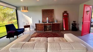 A modern living room with a large window, a sofa, a red door, and a wooden cabinet with decorative items. - Casa Melipill n BBQ Jacuzzi Mini-pool Pet Wifi (Melipilla)