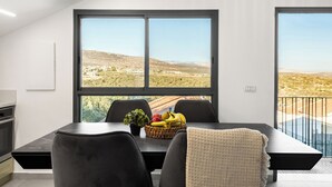 Ein moderner Essbereich mit großem Fenster, einem Esstisch mit Stühlen, einer Obstschale und Blick auf eine hügelige Landschaft.