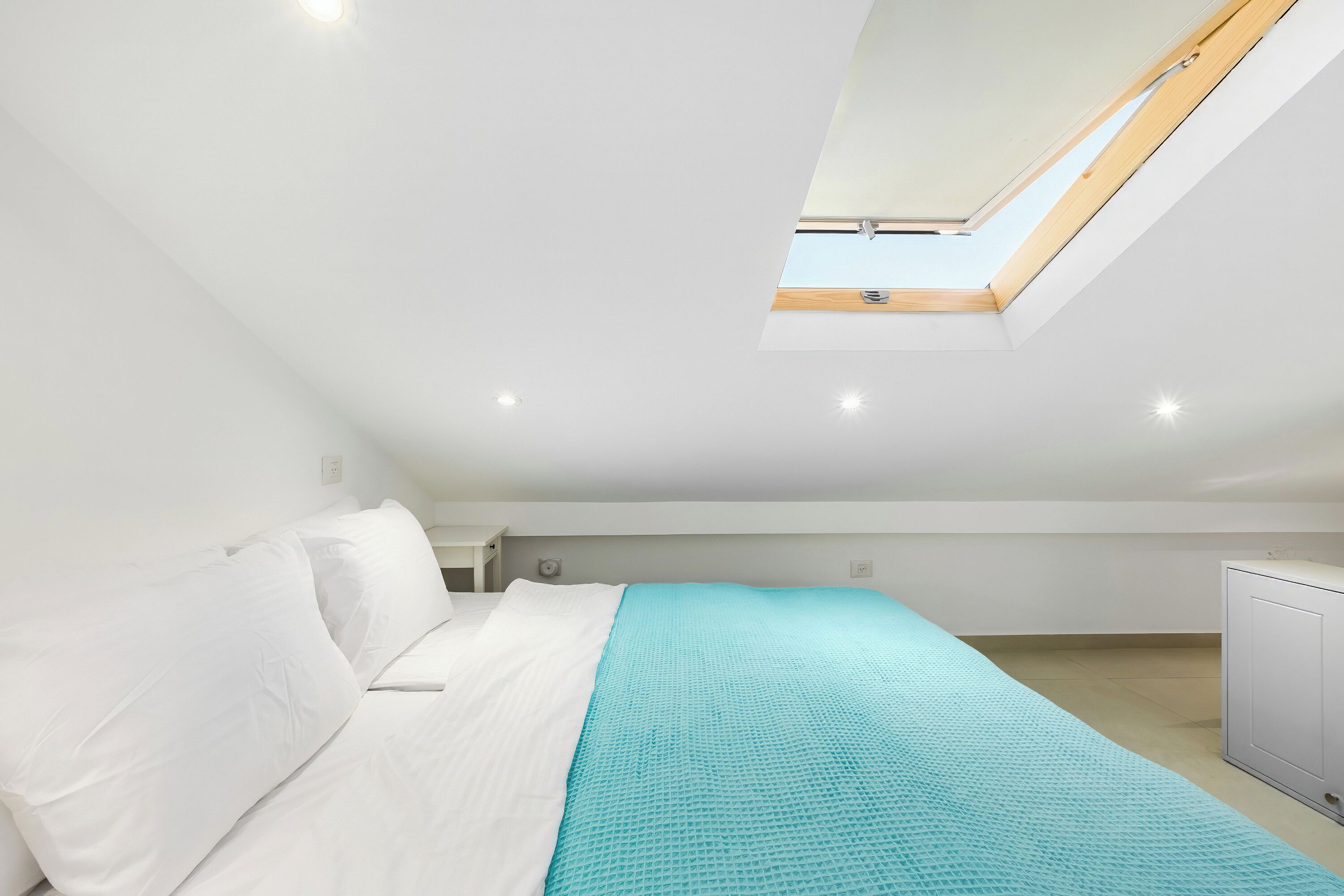 A bedroom with a skylight, a bed with white linens, a teal bedspread, and a white wardrobe.