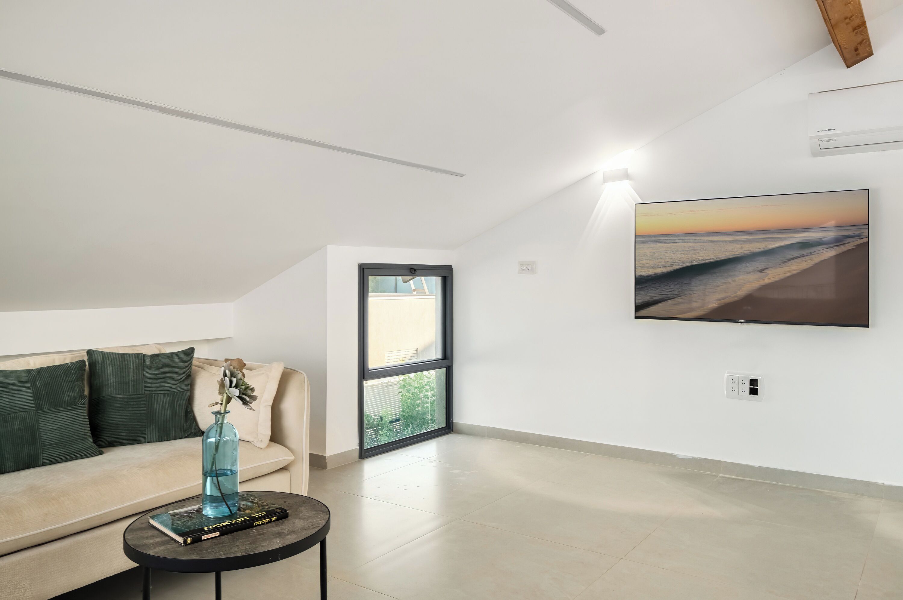 Un salon moderne avec un canapé beige, une petite table ronde sur laquelle sont posés un livre et un vase, une grande fenêtre donnant sur la plage et un tableau représentant la plage accroché au mur.