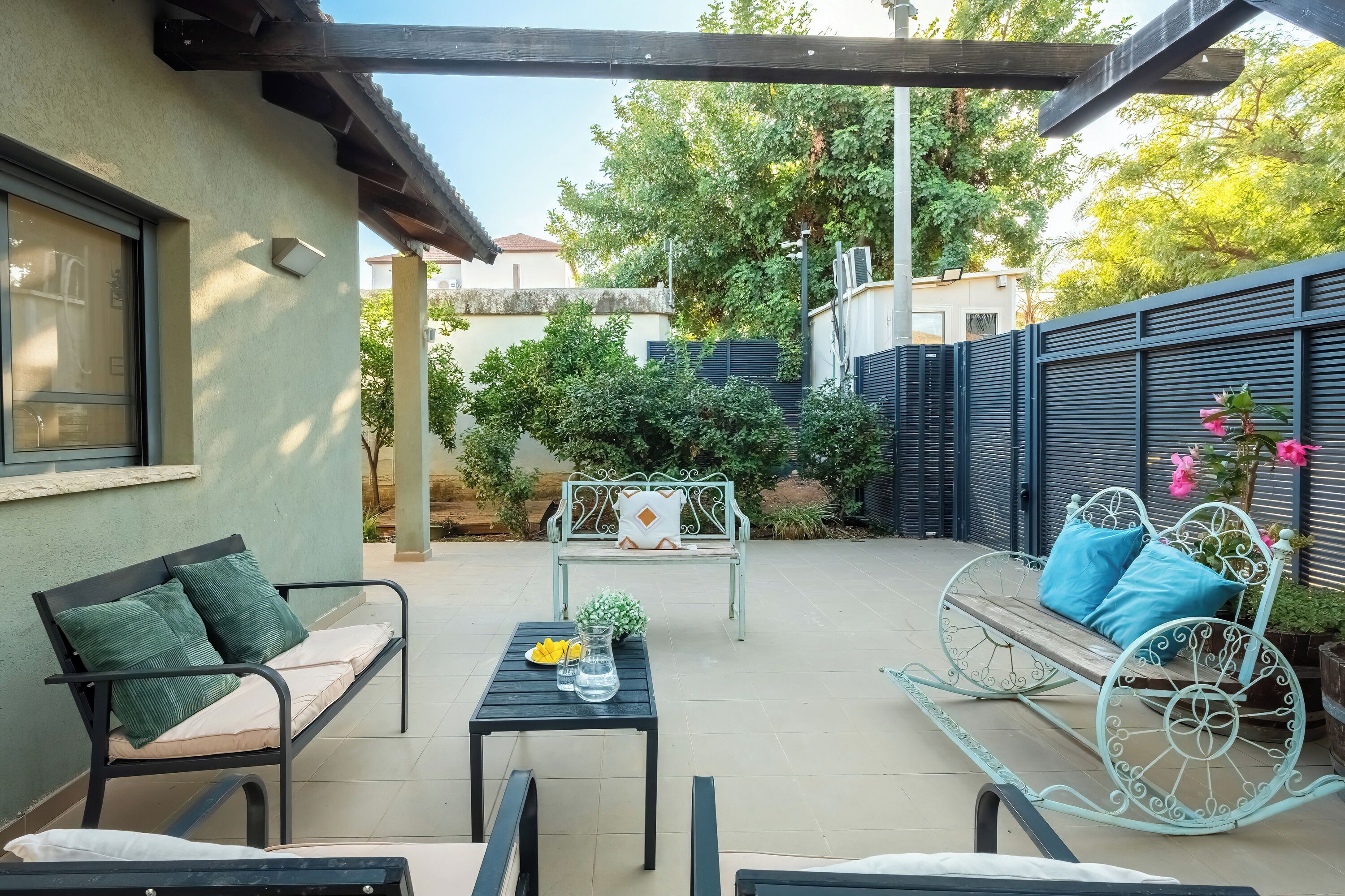 A patio with a metal bench, a swing, and a table with a pitcher and fruit.