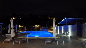 En udendørs swimmingpool med liggestole og parasoller, omgivet af en stenvæg og et overdækket område med et blåt lysloft.