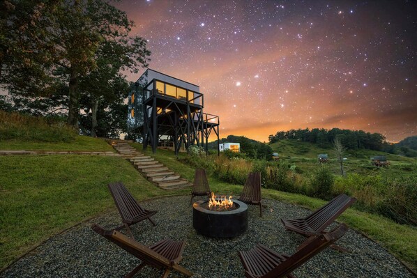 Een sterrenhemel, een vuurplaats met stoelen en een modern huis met een glazen balkon.