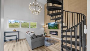 Una sala de estar moderna con un sofá gris, una mesita, una araña, una escalera espiral negra y una chimenea.