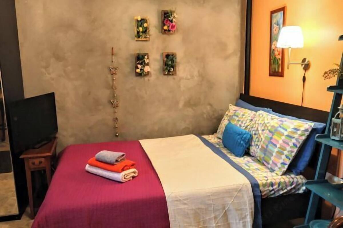 A neatly made bed with a red bedspread and a white blanket, a blue pillow, and a colorful quilt. There are four small wall decorations with flowers and a lamp on a bedside table.